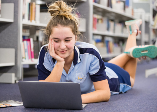 Female high school student in school uniform lying on library floor looking at her laptop