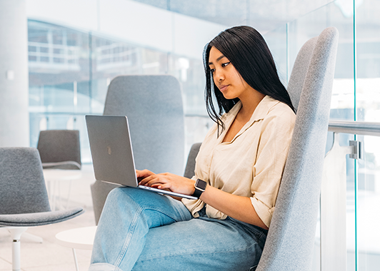 Uni student sitting inside in front of glass windows and typing on laptop