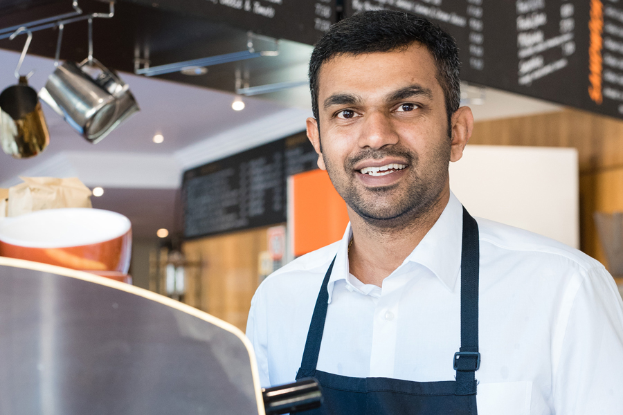 Barista wearing uniform standing in front of coffee machine in a cafe facing camera and smiling